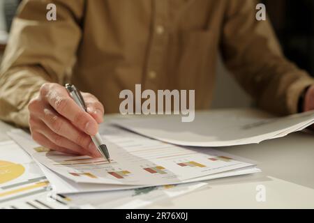 Hand of young economist with pen pointing at financial document with graphic data while analyzing it and preparing report for seminar Stock Photo