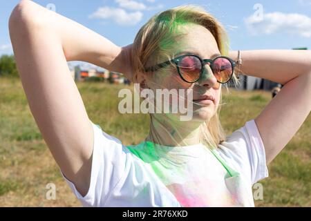 Closeup Beautiful Caucasian Blond Woman With Colorful Dye, Powder On Face, Cloth On Holi Colors Festival In Park, Sunny Day. Playful Cultural Event Stock Photo