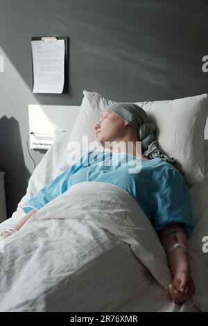 Young woman in headscarf and blue shirt lying in bed in clinics and sleeping during medical procedure while having course of chemotherapy Stock Photo