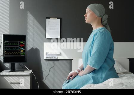 Young recovering female patient of oncological clinics in headscarf and blue uniform sitting on bed in ward and looking through window Stock Photo