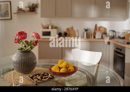 Several yellow pears on plate, nuts and vase with bunch of red flowers on glass round table standing in the center of spacious kitchen Stock Photo