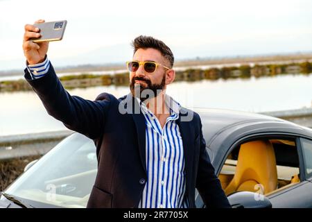 A young male wearing sunglasses taking a selfie while standing next to his sports car Stock Photo
