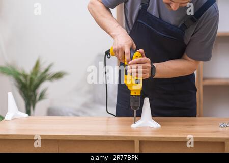 Experienced furniture assembler using an electric screwdriver to attach legs to a wooden rack  Stock Photo