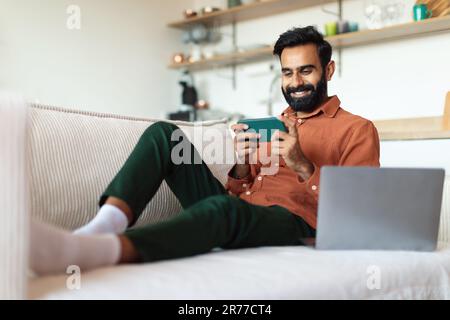 Bearded Indian Man Using Smartphone Sitting Near Laptop At Home Stock Photo