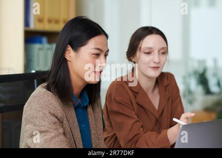 Two young colleagues pointing at monitor and discussing presentation on laptop during teamwork in office Stock Photo