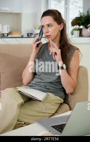 Vertical image of young woman talking on mobile phone while sitting with her notepad on sofa, she working at home Stock Photo