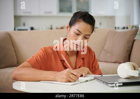 Young woman making notes in copybook sitting at table with laptop, she studying at home Stock Photo
