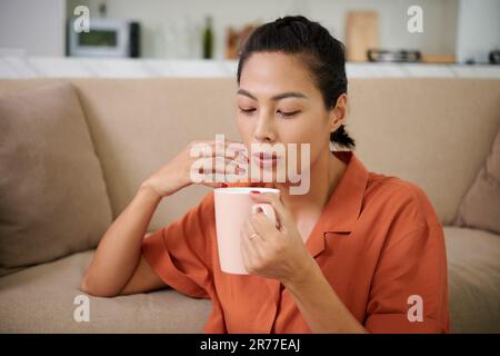 Young woman blowing on cup and drinking hot coffee while resting at home Stock Photo