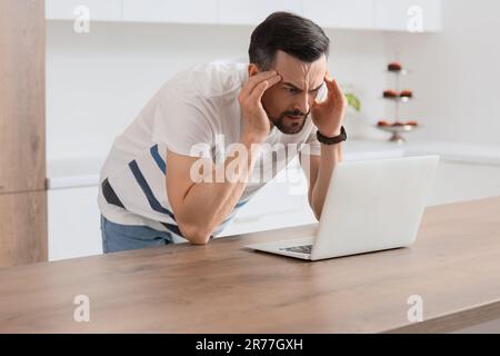 Young man with laptop having panic attack in kitchen Stock Photo