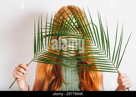 Studio shot of adorable red-haired preteen kid girl, wearing khaki dress, hiding behind exotic green palm leaves Stock Photo