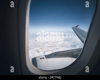 Flying over snow capped mountains. View of snowy landscape under the plane wing through plane porthole. White snow covered space and bright blue sky. Stock Photo