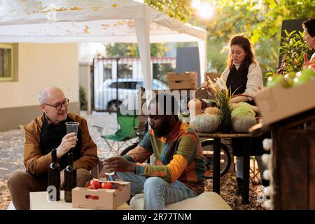 Young African American guy customer tasting homemade natural wine while sitting at table with winemaker at farmers market. Consumer sampling local produce while visiting organic food festival Stock Photo