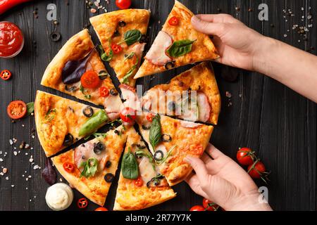 Hands take slices of cut pizza on a dark wooden background top view. Pizza with salami, jerky, sauces, spices, fresh cherry tomatoes on a black Stock Photo
