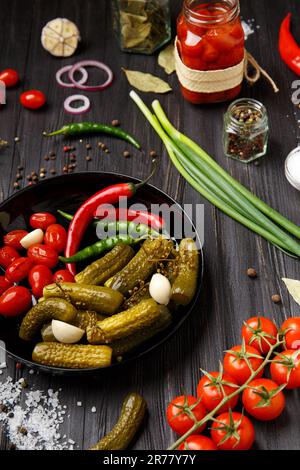 Canned cherry tomatoes, cucumbers, chili peppers in a round black plate on a dark background, close-up. Mix of pickled vegetables, spices, vegetarian Stock Photo