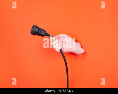 A man's hand holds a black power cable through torn orange paper. Hand through torn orange paper. Stock Photo