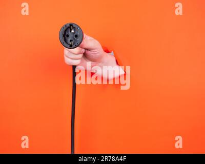 A man's hand holds a black power cable through torn orange paper. Hand through torn orange paper. Stock Photo