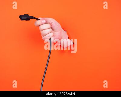 A man's hand holds a black power cable through torn orange paper. Hand through torn orange paper. Stock Photo