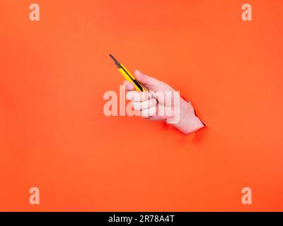 A man's hand holds a yellow cutter through torn orange paper. Hand with sharp knife. Stock Photo