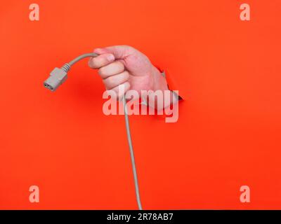A man's hand holds a gray power cable through torn orange paper. Hand through torn orange paper. Stock Photo