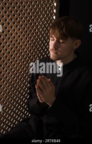 Catholic priest praying near wooden partition in confessional Stock Photo