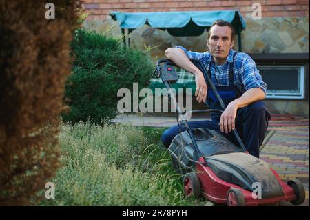 Professional male gardener in gardening uniform, sitting near electric lawn mower, smiling looking confidently at camera Stock Photo