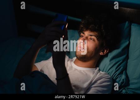 Insomnia, addiction concept. Sleepy exhausted man lying in bed using smartphone, can not sleep. Handsome guy in bed scrolling through social networks Stock Photo