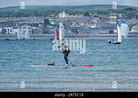Man paddles, standing on paddleboard in Mount’s Bay. Small sailing boats and town of Marazion in background. West Cornwall, England, UK. Stock Photo