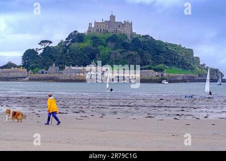 Female walks her dog on Marazion Beach. St Michael’s Mount across Mount’s Bay with sailing boats in the water. Cornwall, England, UK. Stock Photo