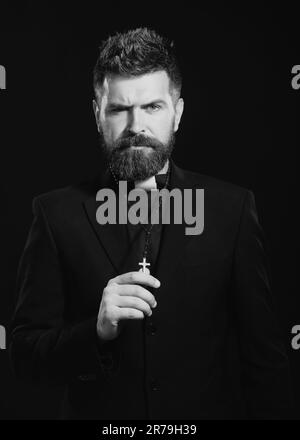 Religion concept. Handsome hispanic catholic priest man over black isolated background. Pastor or preacher praying, priest portrait of male pastor. Stock Photo