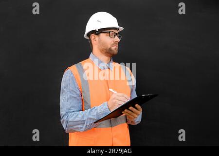 Male Engineer in a White Hard Hats and Orange West. Handsome Confident Build Worker Isolated on Blank Black Background. Copy Space. He Work in a Heavy Stock Photo
