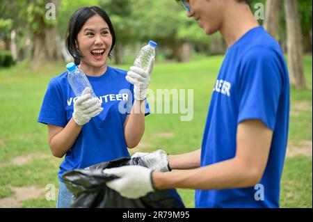 A joyful and happy young Asian female volunteer collecting trash and plastic bottles into a garbage bag with her friend, helping their community by cl Stock Photo