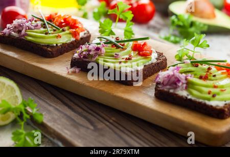 Fresh vegetarian open sandwiches on black bread with avocado, tomato and red onion on the wooden board. Concept healthy eating Stock Photo