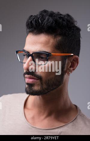Side portrait of a fit, attractive Middle Eastern man. Black hair, neat beard, beige shirt, colorful plastic glasses, and an earring highlight his app Stock Photo