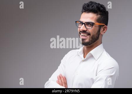A handsome man laughs with crossed arms. He wears a white shirt, black and orange glasses, on a grey background Stock Photo