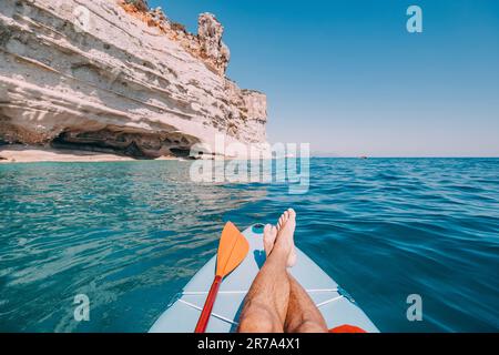 Men's legs and paddle on a supboard against the backdrop of a picturesque seascape with a rock. Relaxation and fitness on the water Stock Photo