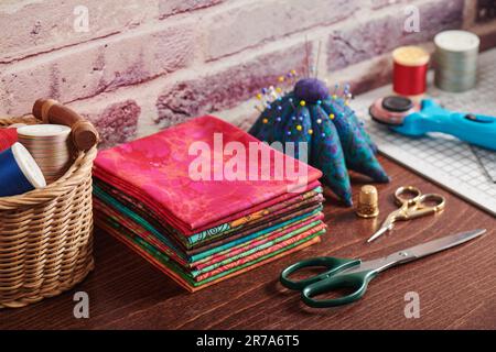 Stack of colorful fabrics on table surrounded by sewing accessories on brick wall background Stock Photo