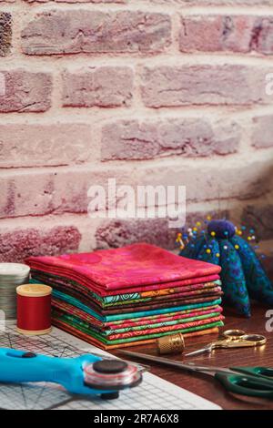 Stack of colorful fabrics on table surrounded by sewing accessories on brick wall background Stock Photo