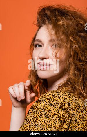 Portrait of young red haired model in yellow blouse with abstract print looking at camera and standing isolated on orange, stylish casual outfit and s Stock Photo
