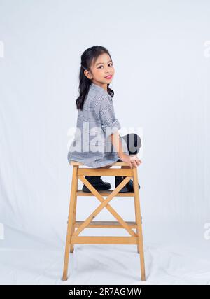 A young woman is seated in a wooden chair, her legs crossed in a relaxed pose Stock Photo