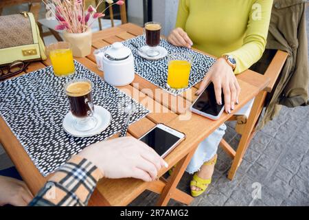 top view of two unrecognizable young women taking the phone from a table in the cafeteria, waiting for the food for breakfast, with cup of coffee and Stock Photo