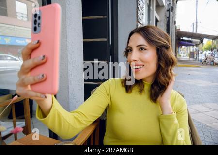 young long-haired latin woman of argentinian ethnicity dressed in a sweater, happy smiling taking a selfie with her phone, sitting outside the coffee Stock Photo