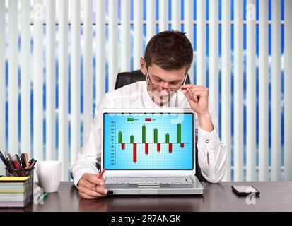 young trader sitting in office and looking on screen laptop with cost and profit chart Stock Photo