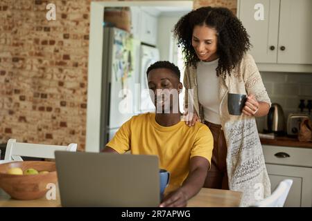 Smiling multiethnic couple using a laptop in their kitchen in the morning Stock Photo