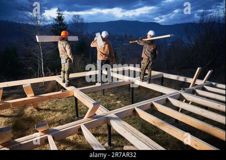 Men workers building wooden frame house on pile foundation. Back view of carpenters carrying wooden truss for timber framing. Carpentry concept. Stock Photo