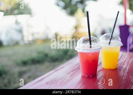 Two bright ice cocktails in plastic cups with straws on a wooden table in an open air cafe. Close up of delicious berry and citrus juices outdoors. Gr Stock Photo