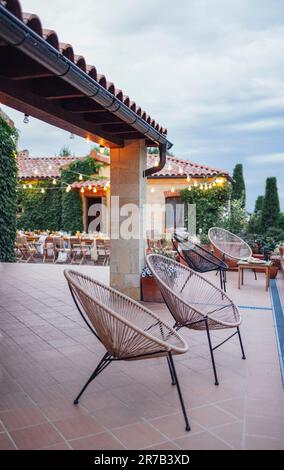 Stylish European outdoor cafe in the evening. Wooden tables and chairs, wicker chairs, flower pots and garlands of light bulbs. Ivy-covered light-bric Stock Photo