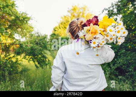 Young woman in casual wear carries huge bouquet of flowers on her shoulder outdoors in meadow. White girl standing back and holding irises and chamomi Stock Photo