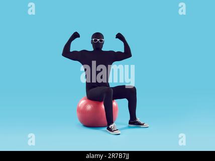 Faceless man in black bodysuit and hands in fists sitting on sport fit ball on blue background. Stock Photo