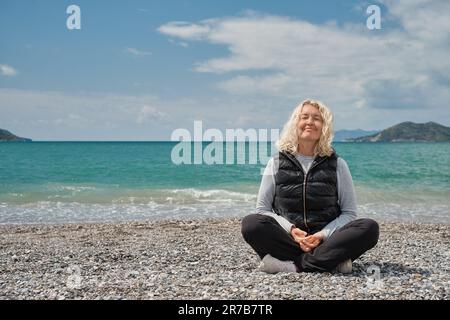 blonde sits on a pebbly beach on a spring day. Outdoor portrait of beautiful curly adult european woman smiling on sea background Stock Photo