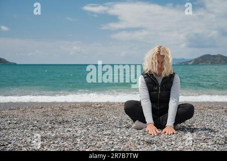 blonde european woman sits on a pebble beach on a spring day touches the stones on the beach with her hands. Outdoor portrait of a beautiful curly adu Stock Photo